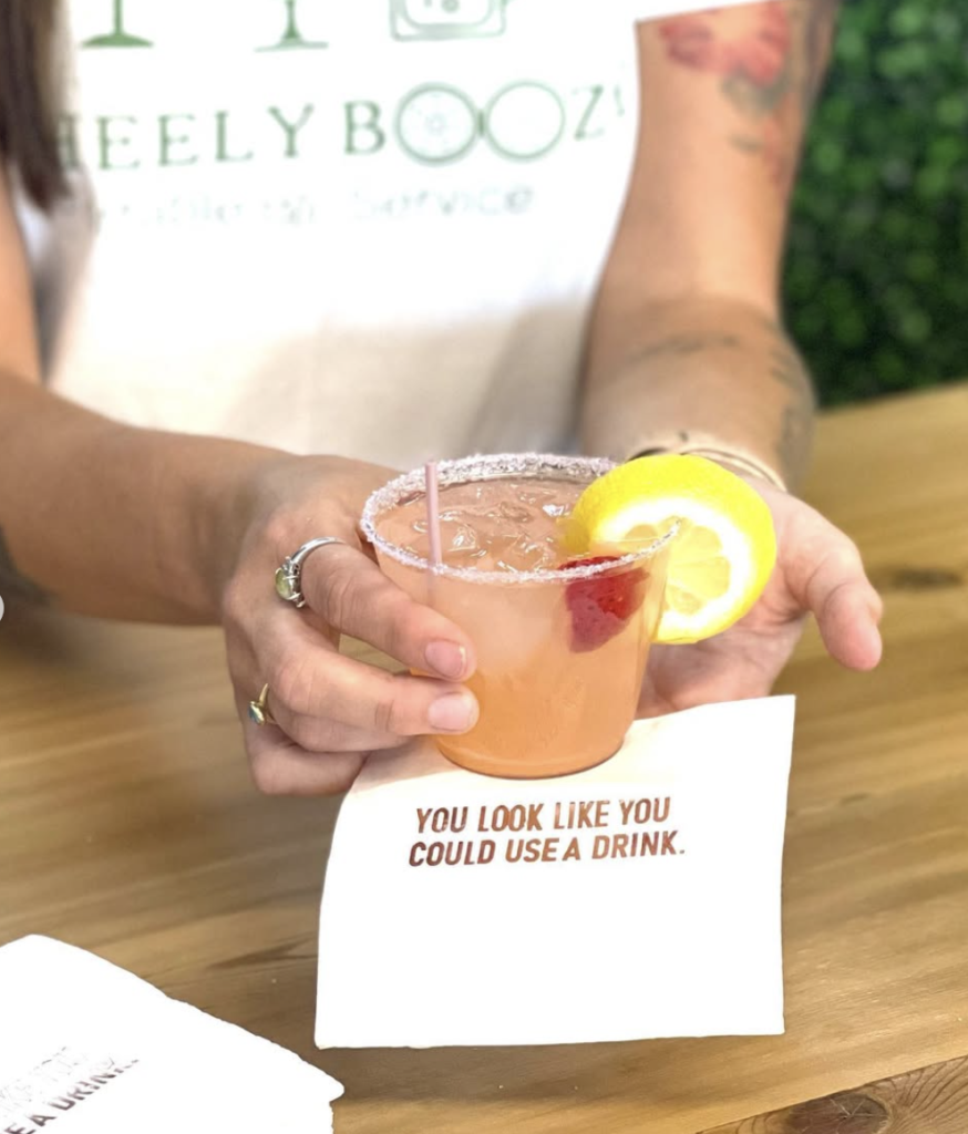 Hands holding pink cocktail garnished with lemon slice and cherry over wooden table with playful napkin