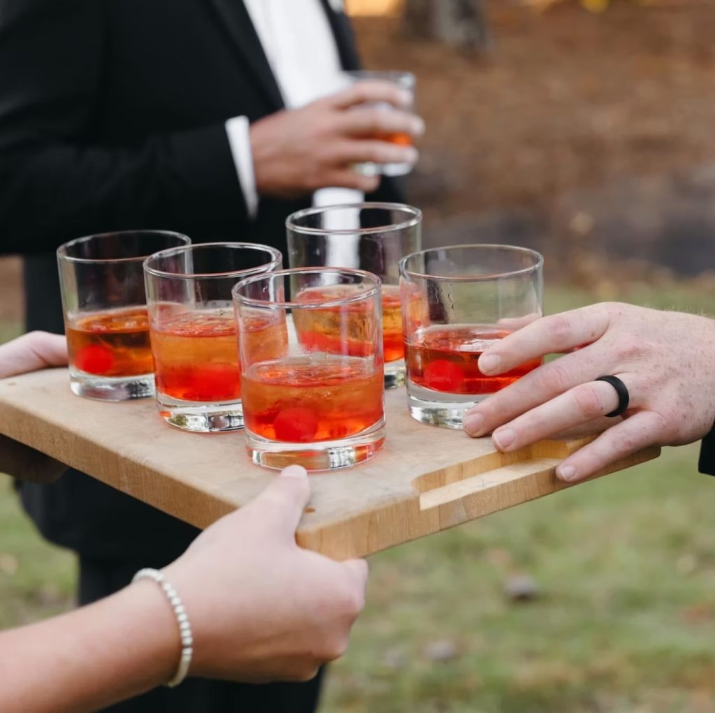 Server presenting red cocktails on wooden tray to wedding guests outdoors