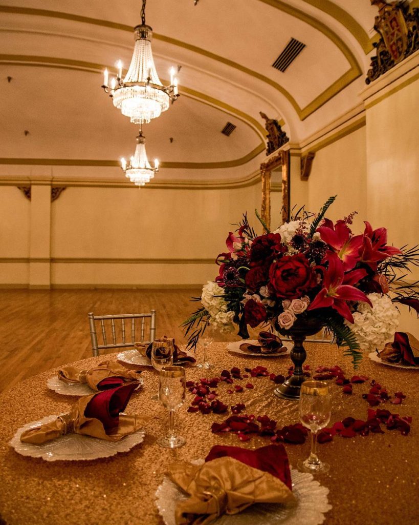 Elegant ballroom table setting with red floral centerpiece, gold linens, and crystal chandeliers