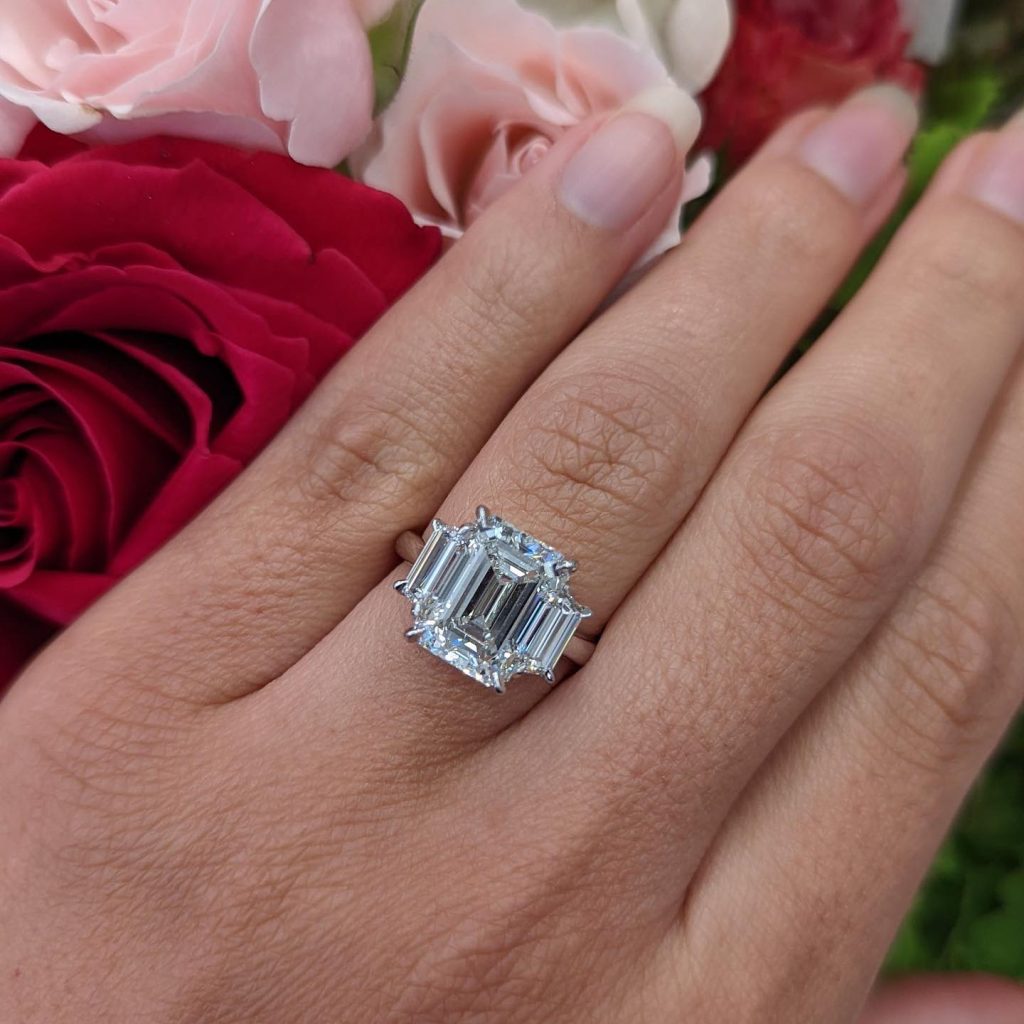 Hand displaying emerald-cut diamond engagement ring with halo setting among red and pink roses