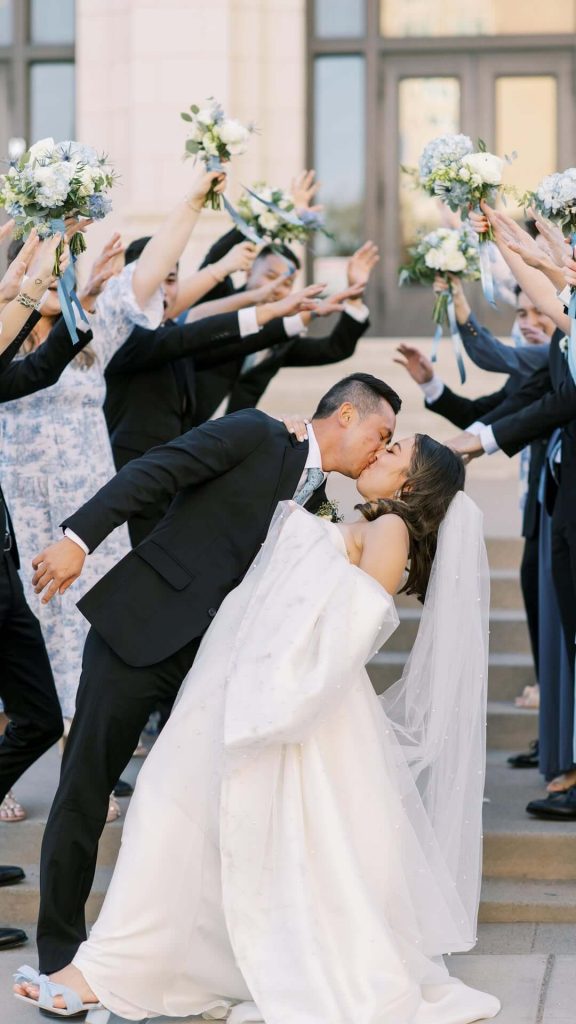 Bride and groom share a dip kiss while wedding party raises bouquets with blue and pink ribbons in celebration