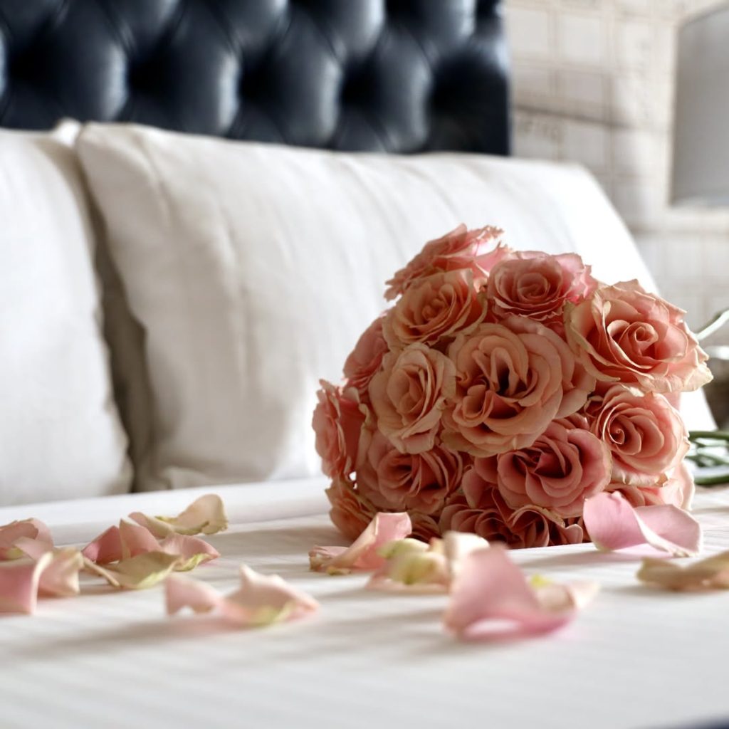 Coral pink rose bridal bouquet on white bed with scattered petals in luxury hotel suite