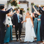 Newlyweds walk under floral arch held by wedding party in navy suits and teal bridesmaid dresses