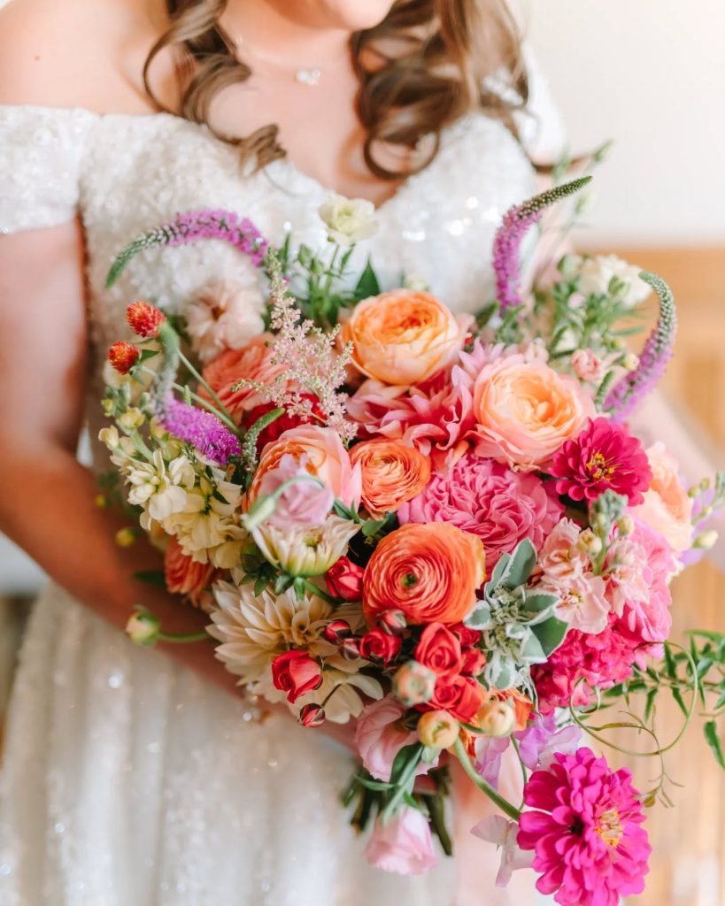 Bride holding vibrant bouquet with peach, pink, and fuchsia blooms in white beaded gown