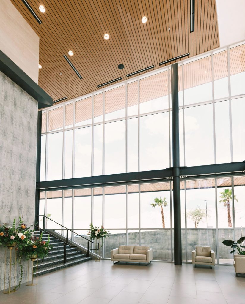 Modern event space lobby with floor-to-ceiling windows, wood-slat ceiling, grand staircase, and neutral lounge furniture