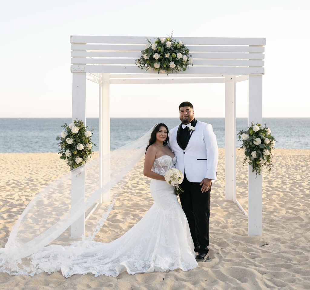 Bride in lace mermaid wedding dress with groom under white arbor decorated with roses on beach
