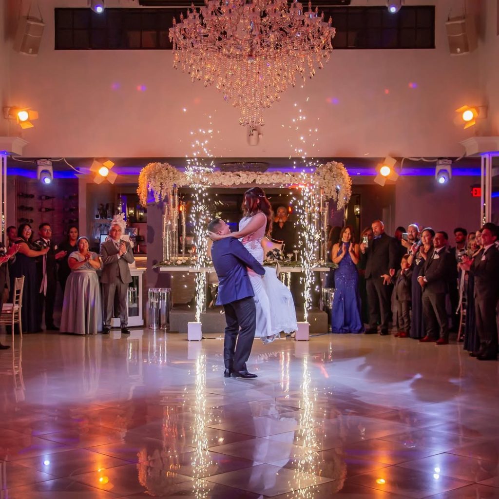 Bride and groom sharing first dance under sparkling chandelier with indoor fireworks and wedding party surrounding dance floor