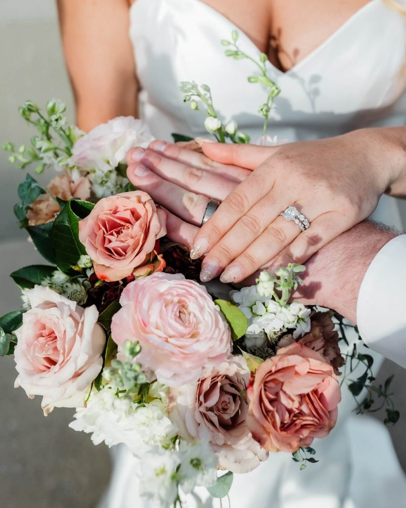 Hands of bride and groom displaying wedding rings over pink rose bouquet