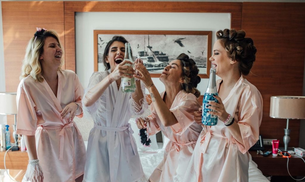 Bride and bridesmaids in pink robes laughing and toasting with champagne during wedding day preparation