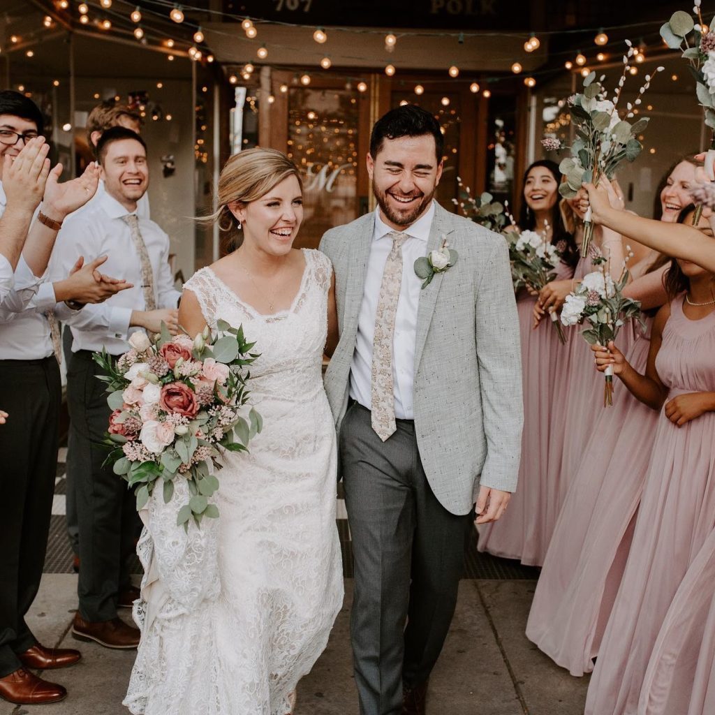 Newlyweds walking down aisle under string lights as wedding party cheers, bride in lace gown holding floral bouquet