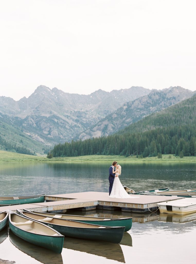 Bride and groom embracing on mountain lake dock surrounded by green canoes with forested peaks in background