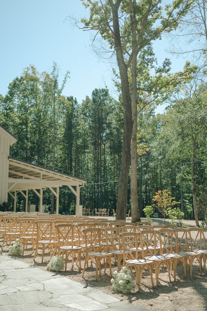 Outdoor wedding ceremony setup with wooden chairs facing rustic pavilion surrounded by trees
