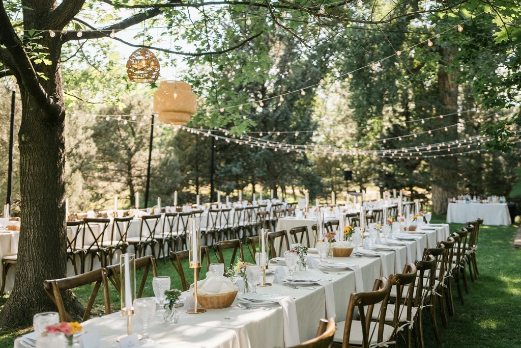 Outdoor wedding reception with long tables, string lights, and hanging lanterns under shaded trees