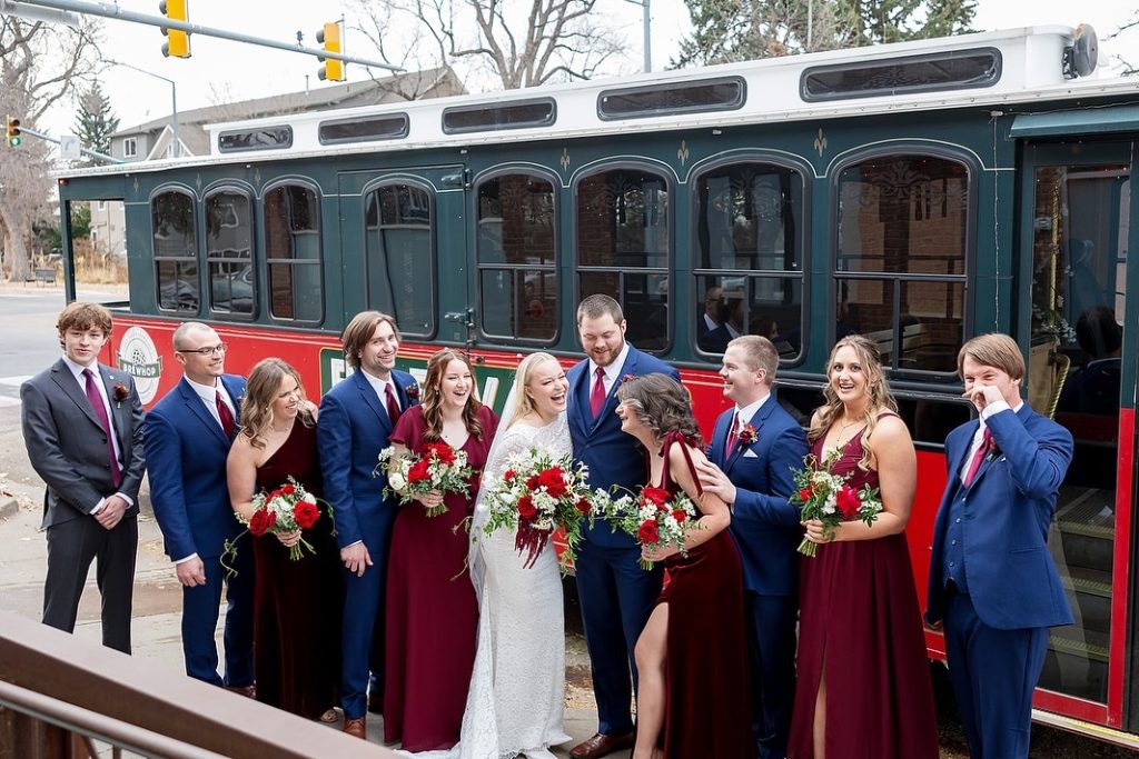 Wedding party in navy suits and burgundy dresses posing in front of vintage green and red Brewhop Trolley