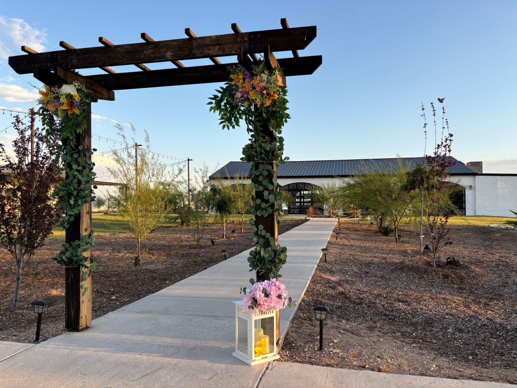 Wooden pergola decorated with colorful floral arrangements framing a walkway leading to Hacienda Sol Y Luna's main venue building
