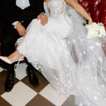 Bride and groom seated on red leather chairs, showcasing floral-detailed wedding dress and white bouquet