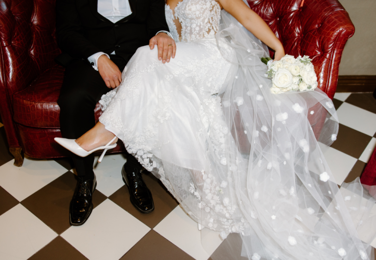 Bride and groom seated on red leather chairs, showcasing floral-detailed wedding dress and white bouquet