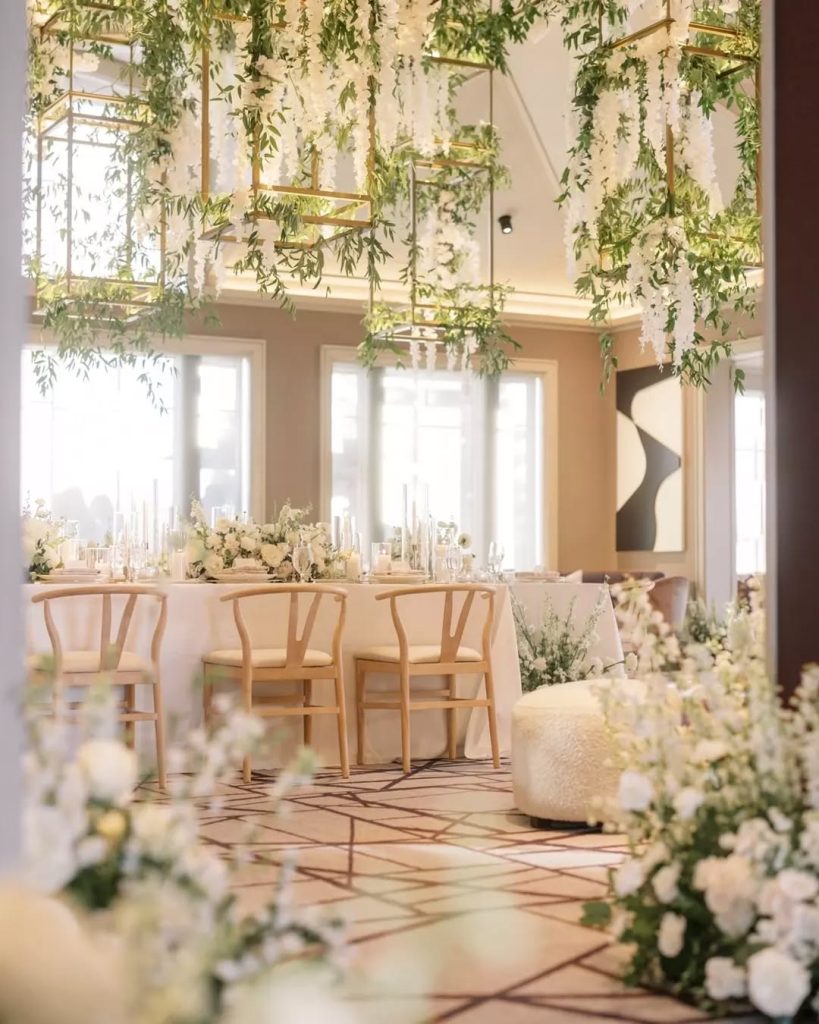 Elegant wedding reception with gold geometric frames adorned with cascading greenery above long tables with modern wooden chairs