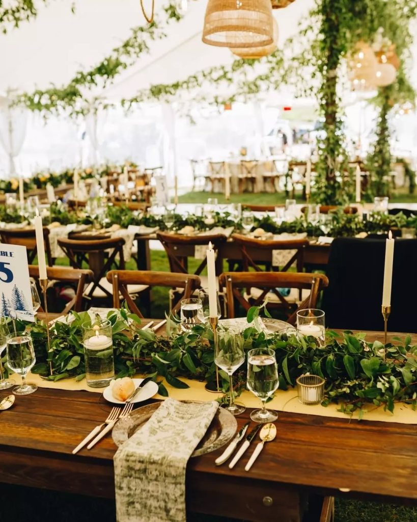 Wooden farm tables decorated with lush greenery garlands, white candles, and rustic cross-back chairs under white tent