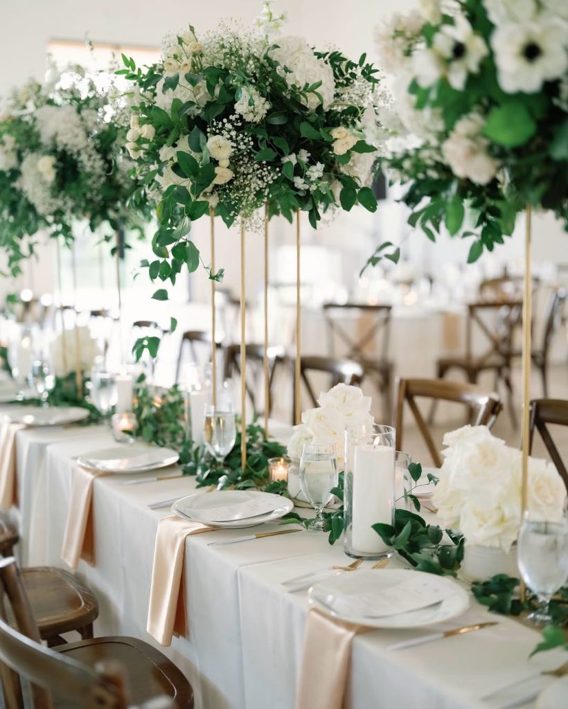 Elegant wedding reception tables with tall gold centerpieces featuring white flowers and greenery, set with neutral linens