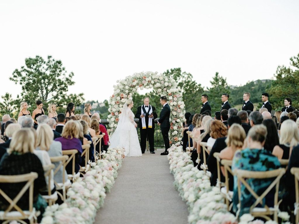Outdoor wedding ceremony with floral arch and aisle lined with white flowers, guests seated watching couple exchange vows