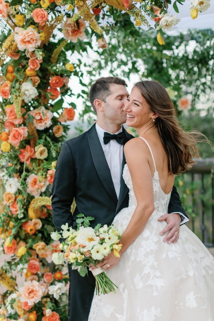 Bride and groom embracing beneath vibrant peach, yellow, and white floral wedding arch at outdoor Denver ceremony