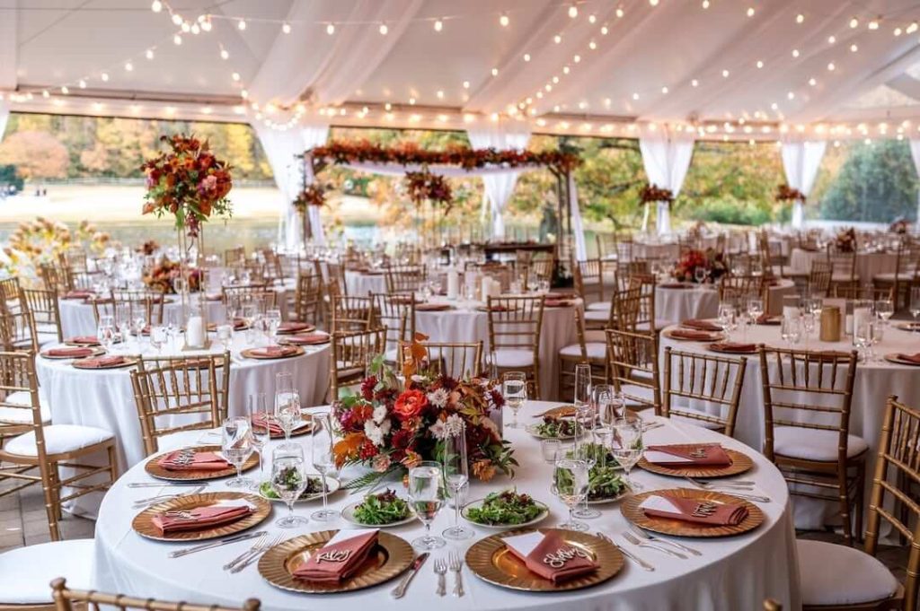 Tented wedding reception with fall-colored floral centerpieces, gold chiavari chairs, and string lighting overhead