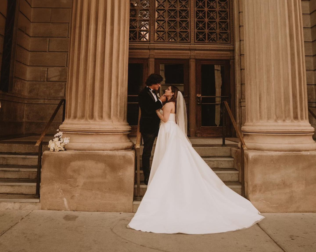 Bride in strapless ballgown wedding dress with cathedral train kissing groom on stone steps beneath classical columns