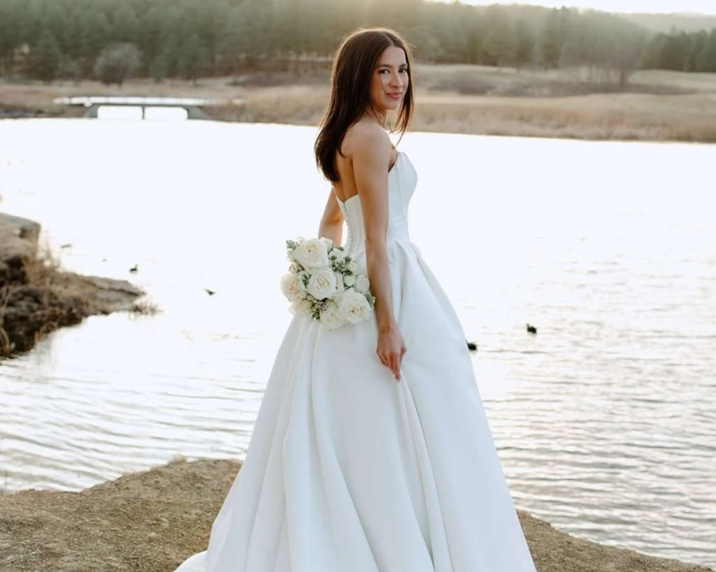 Bride in classic white ball gown holding white rose bouquet beside lake at sunset