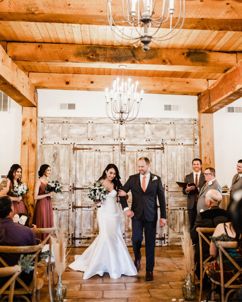 Newlyweds walking down ceremony aisle in rustic barn venue with exposed wood beams, white chandelier, and weathered door backdrop
