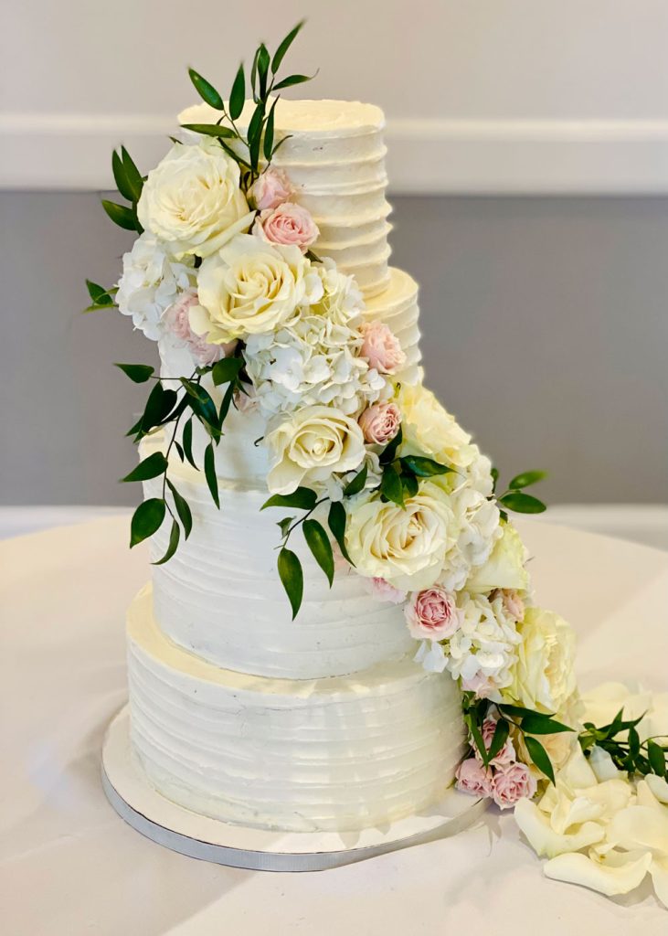 Four-tier white wedding cake with textured buttercream and cascading fresh flowers including ivory roses, pink spray roses, white hydrangeas, and greenery