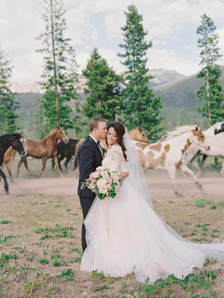 Bride and groom embracing in mountain meadow with herd of horses running behind them beneath evergreen trees