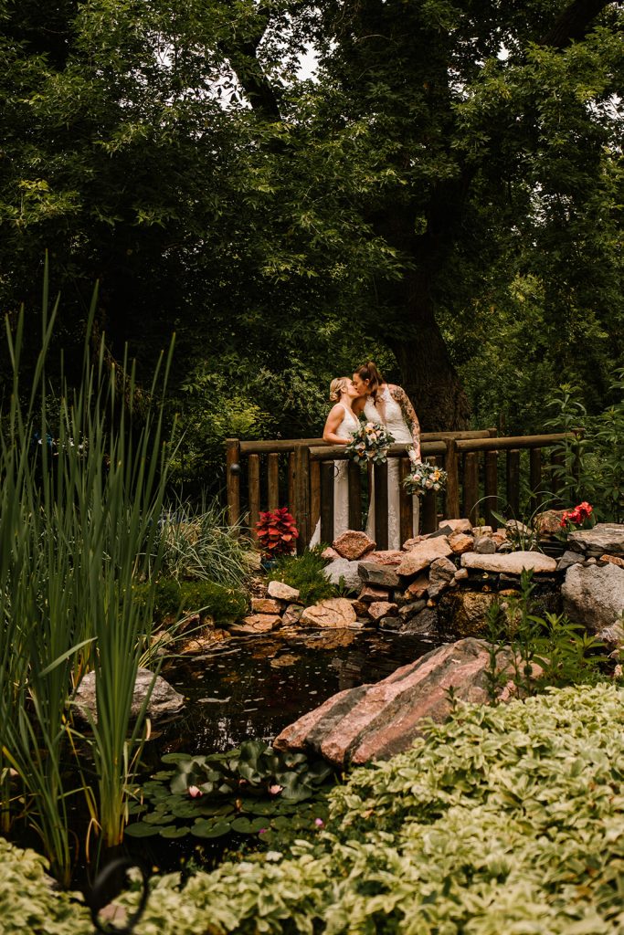 Two brides kissing on a rustic wooden bridge over a lily pond surrounded by lush garden foliage