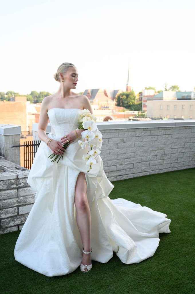 Bride in strapless gown holding white orchid bouquet on Raleigh rooftop with city skyline behind