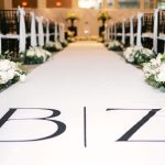 Wedding ceremony aisle with white runner featuring couple's initials, lined with white floral arrangements and gold accents