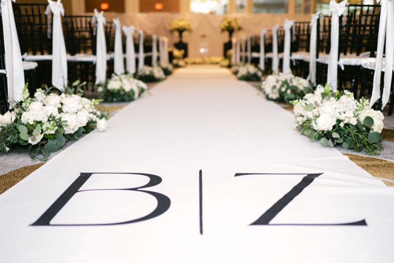 Wedding ceremony aisle with white runner featuring couple's initials, lined with white floral arrangements and gold accents