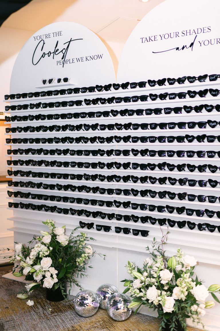 Wedding sunglasses display wall with personalized black sunglasses, white floral arrangements, and disco balls at base