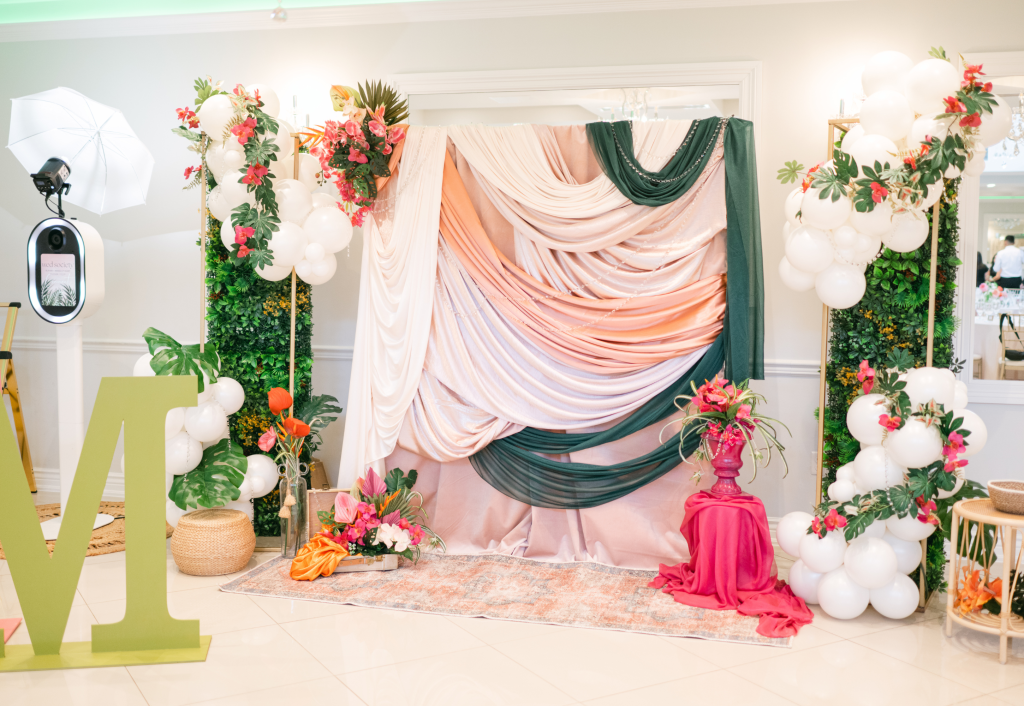 Elegant photo booth setup with blush and emerald draped fabric backdrop, white balloon garland with coral flowers, greenery wall, and tropical floral arrangements