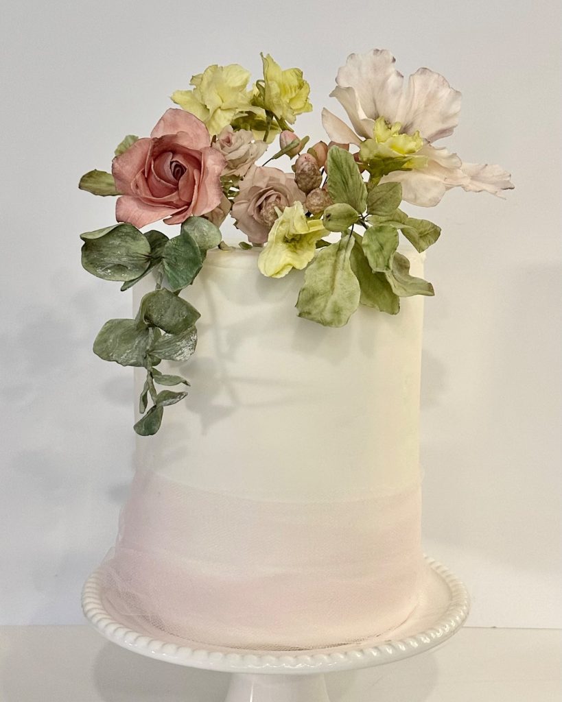 Two-tier white wedding cake with cascading sugar flowers in blush pink, cream, and yellow with eucalyptus accents