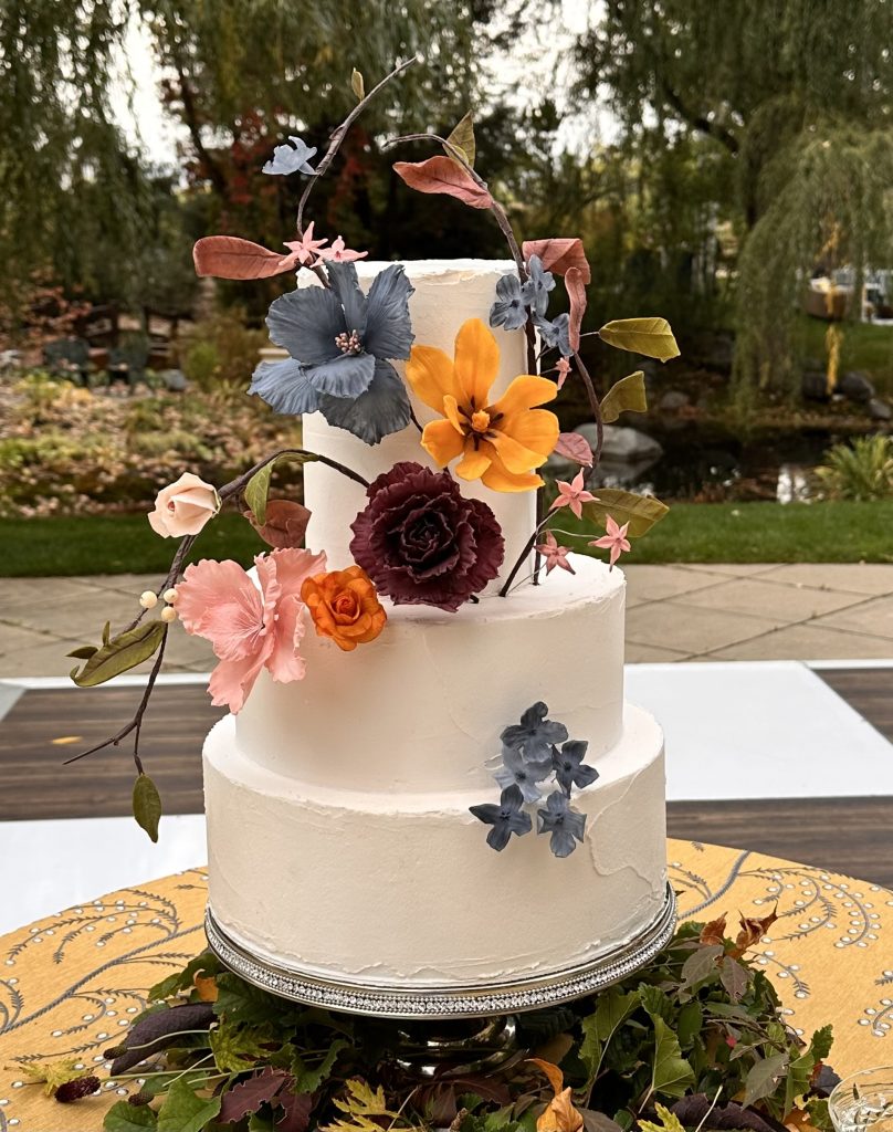 Two-tier white buttercream cake decorated with handcrafted sugar flowers in blue, orange, burgundy, and pink