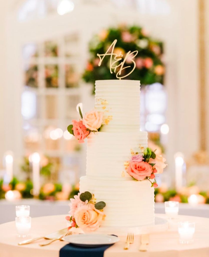 Three-tier white wedding cake decorated with pink blooms and gold monogram topper