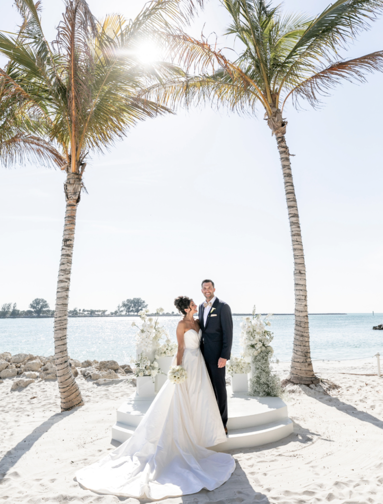 Bride and groom on Clearwater Beach between palm trees with white floral arrangements and Gulf of Mexico backdrop