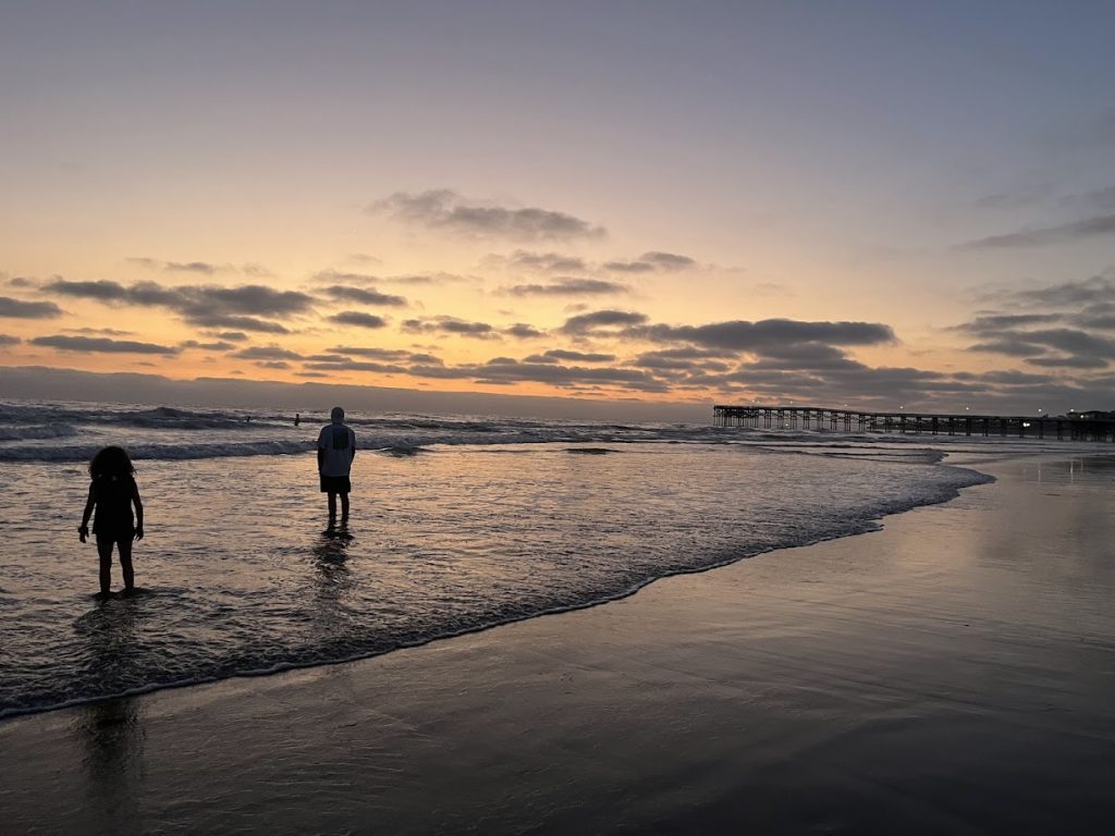 Couple walking along beach at sunset with pier in distance and golden clouds overhead
