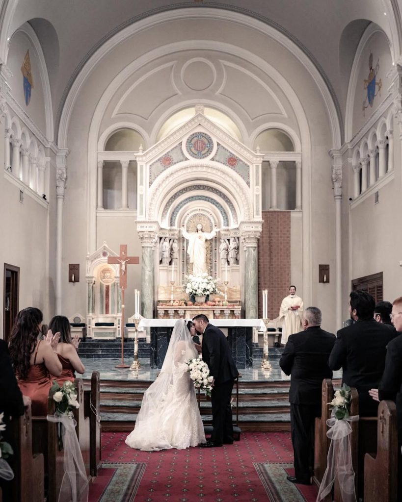 Bride and groom exchanging vows at ornate Catholic church altar with white arched architecture and religious statuary