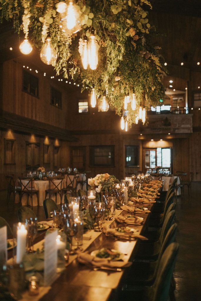 Elegant barn reception hall with long candlelit tables, hanging greenery chandelier, and Edison bulb lighting