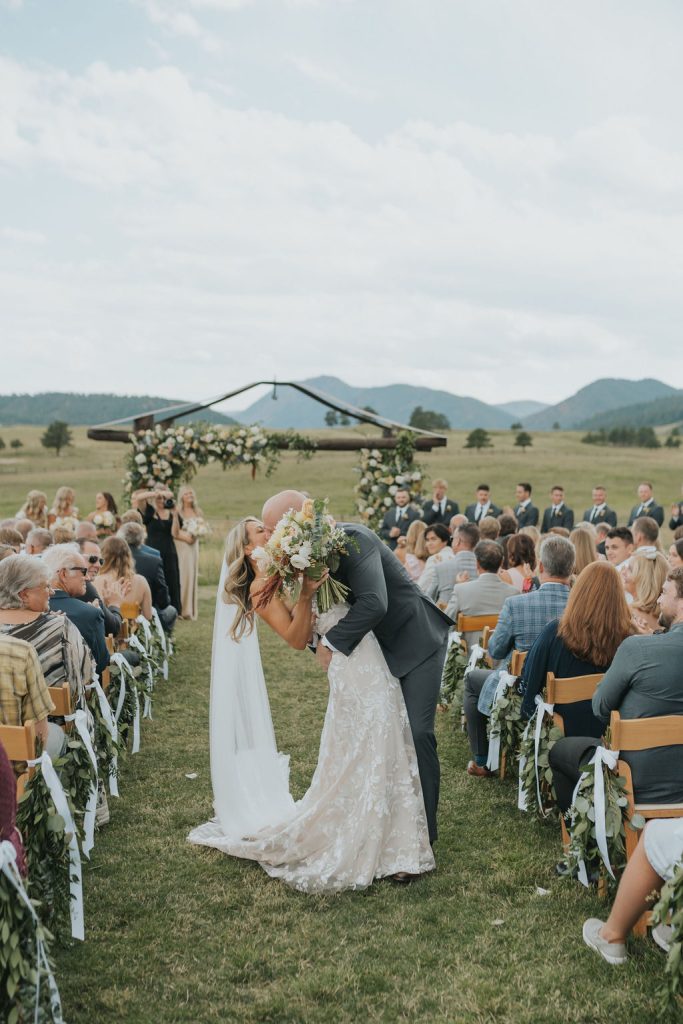 Outdoor ranch wedding with bride and groom kissing under ceremonial arch surrounded by seated guests