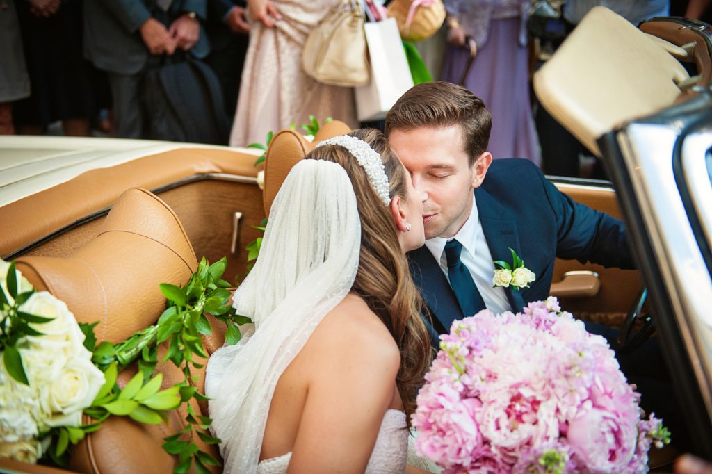 Bride and groom sharing an intimate kiss in vintage car surrounded by wedding party