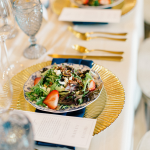 Wedding reception place setting with fresh strawberry salad on gold charger, navy napkin, and gold flatware
