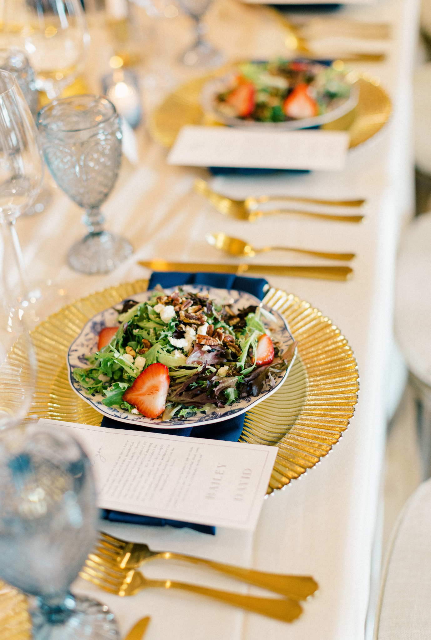 Wedding reception place setting with fresh strawberry salad on gold charger, navy napkin, and gold flatware