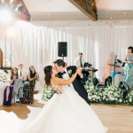 Bride and groom first dance in rustic wedding venue with exposed beams and greenery wall